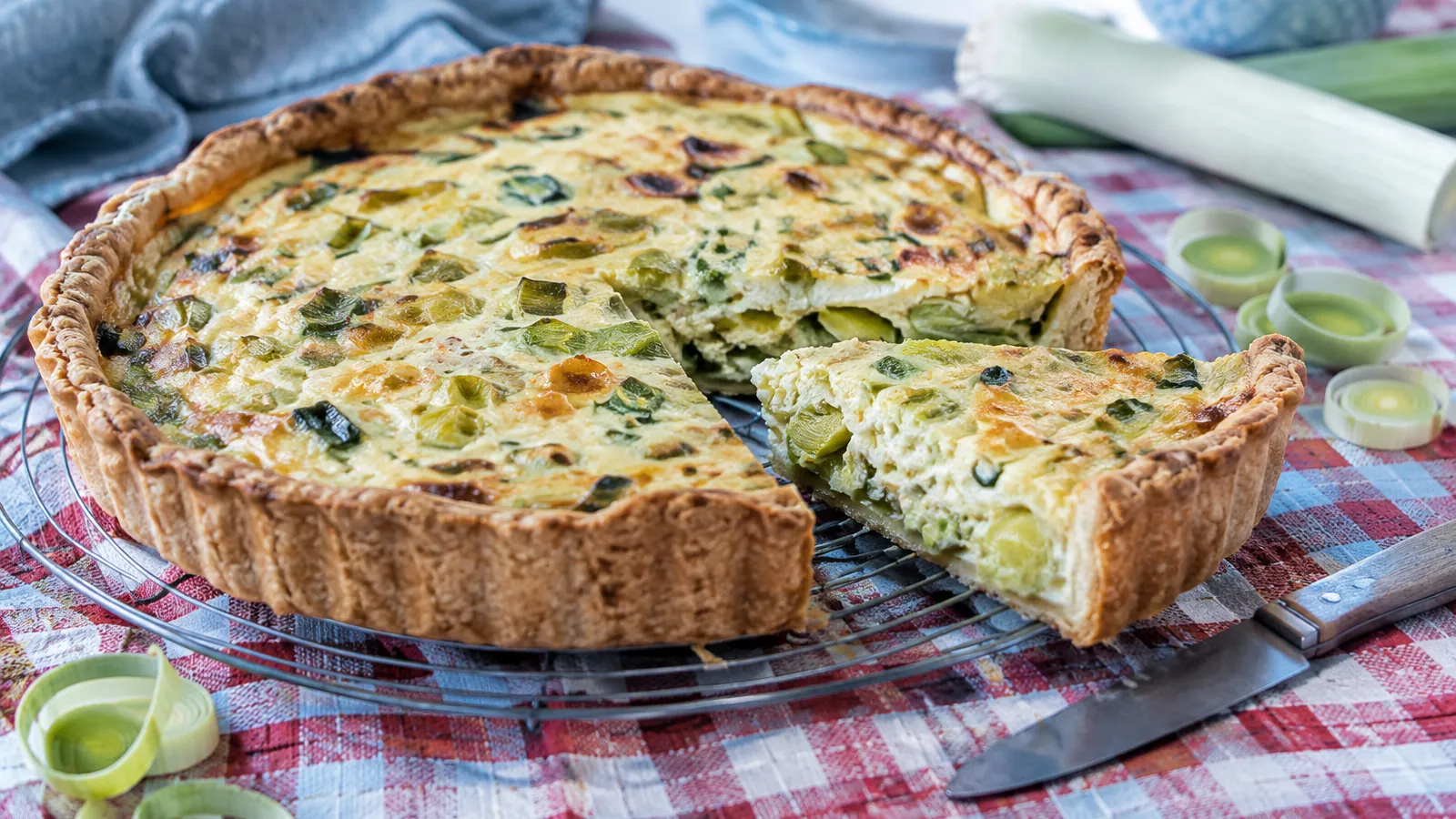 Tarte aux poireaux dorée sur une grille, une part découpée montrant la fondue, nappe à carreaux