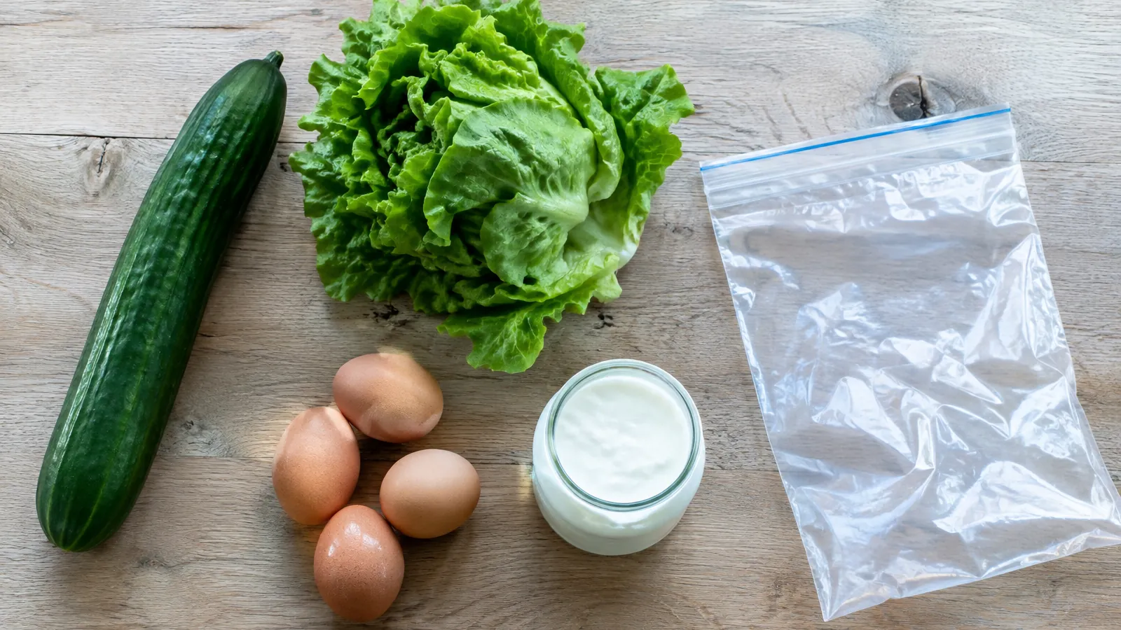 Concombre, salade verte, œufs et pot de yaourt sur une table en bois à côté d'un sachet de congélation vide