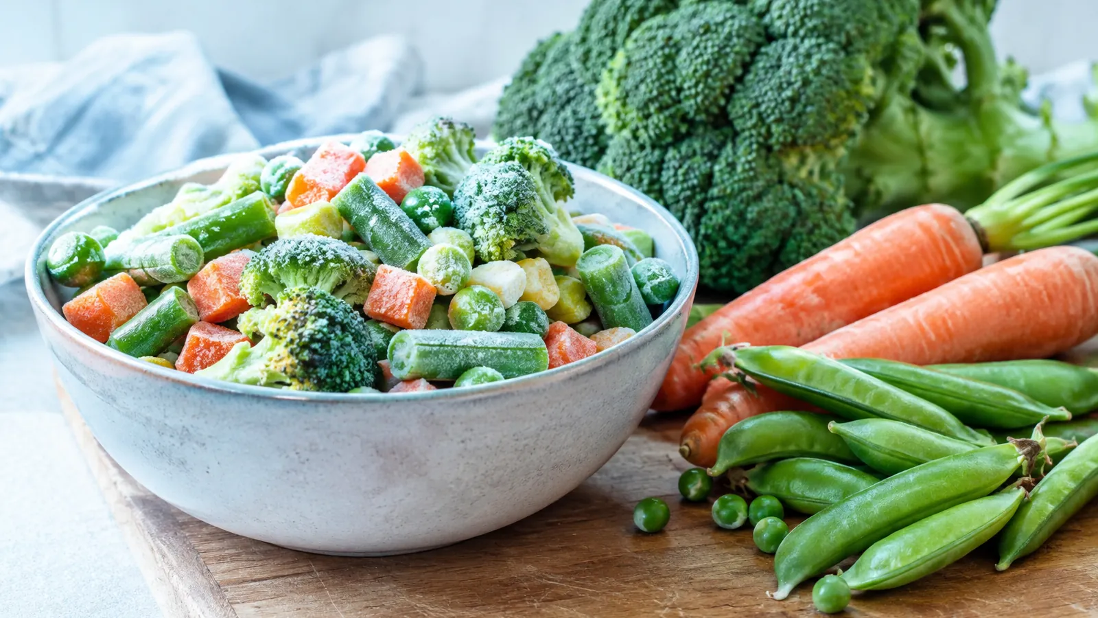 Bol de légumes surgelés givrés à côté de légumes frais sur une planche en bois, cuisine lumineuse